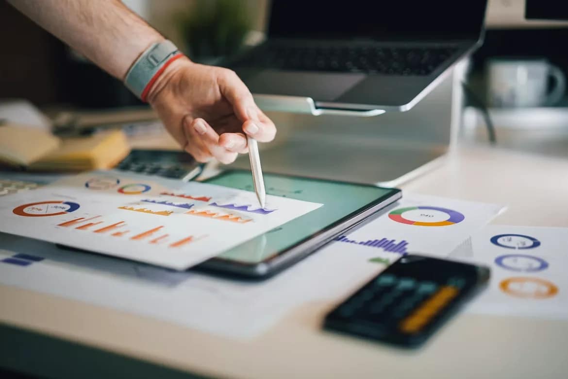 Hand wijst met een pen naar grafieken op papier en een tablet op een bureau